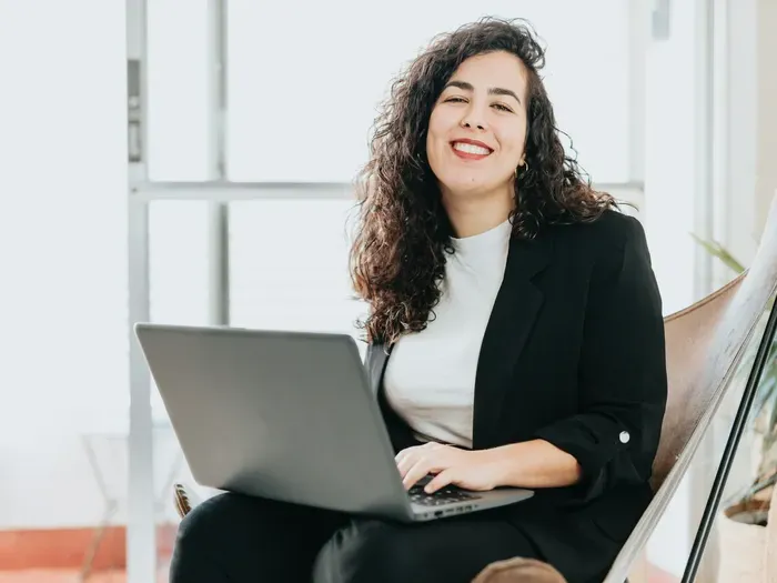 Confident woman in business attire smiling while working on a laptop in a bright modern office setting
